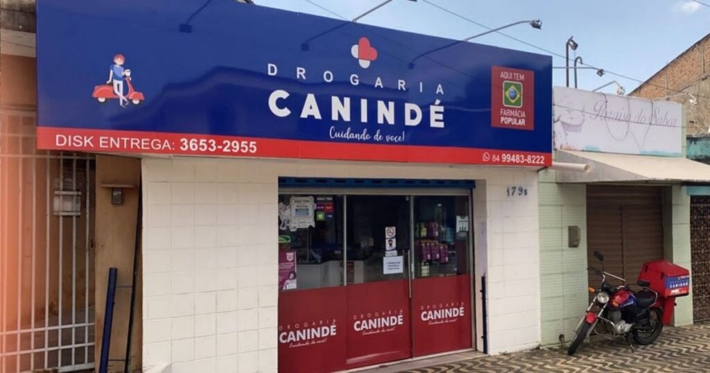 Fachada da Drogaria Canindé com letreiro azul, loja aberta e números de telefone para entrega.